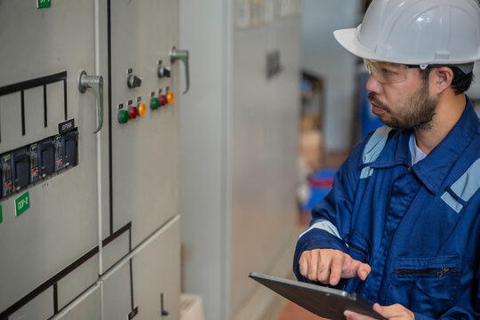 Focused professional male industrial electrical engineer safely uses a digital tablet to inspect a large power control panel during maintenance inside a modern factory manufacturing plant.