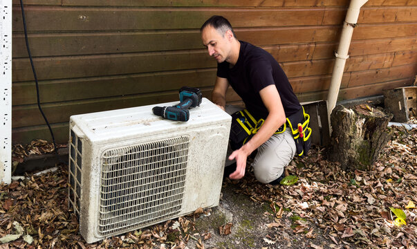 HVAC technician services an outdoor unit near a house wall, holding a tool. Concept of home heating, cooling and maintenance.
