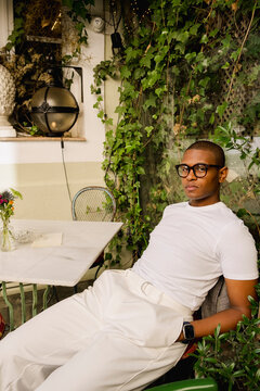 Man in white outfit posing at leafy outdoor cafe table editorial