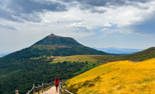 blonde woman hiking on wooden walkway in green forested hills with rope railings, serene mountain landscape overhead, peaceful nature adventure travel scene, The Puy de Dome in Auvergne