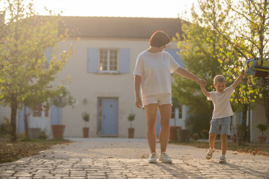 Mother and child walking hand in hand, in cobbled path house with blue shutters trees, with cobblestones, house, blue shutters, trees, from a eye-level perspective perspective.
