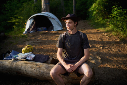 Young Man Sits on Log Near Campsite in Forest During Daytime