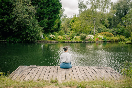 Person Sitting on a Dock by a Calm River Surrounded by Trees