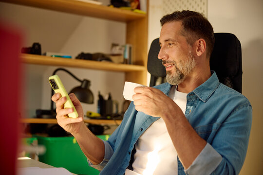 Man sits in the home office, drinking coffee and using his phone