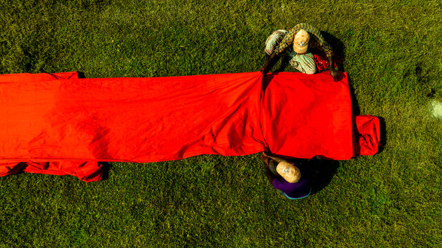 Narsingdi, Bangladesh - 19 November 2022: Aerial view of two people working with a large piece of red fabric on a green grassy field.