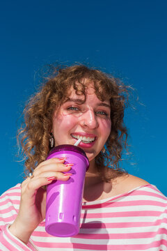 Smiling woman drinking from purple tumbler under blue sky
