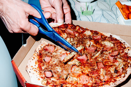 a close-up of a man cutting a delivery pizza with kitchen scissors