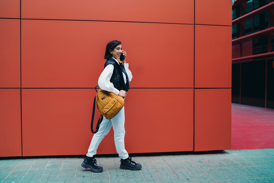 Hispanic woman calling phone mobile roaming connection conversation while walking. Spanish female student traveler with backpack text chatting on cellular near bright red color wall background.