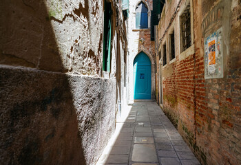 Obraz premium Venice, Italy - May 19, 2026: Narrow cobblestone alleyway in Venice, Italy, featuring a vibrant turquoise door and textured brick and stone walls