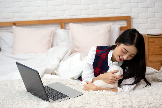Smiling woman cuddling small white Maltese dog beside laptop on bed, showing pet companionship, affection, home comfort, remote work break, relaxation and caring lifestyle
