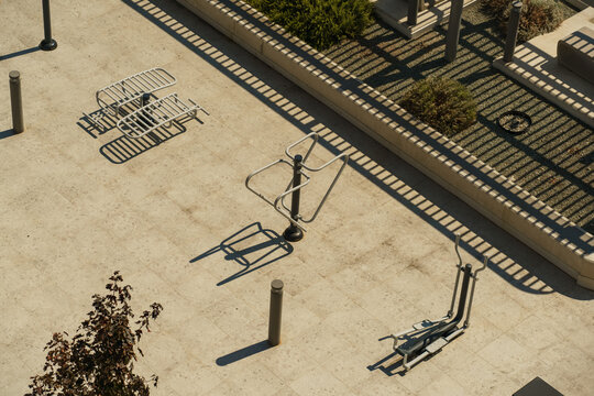 Modern outdoor fitness equipment arranged on a paved urban recreation area, captured from above with strong sunlight creating geometric shadows