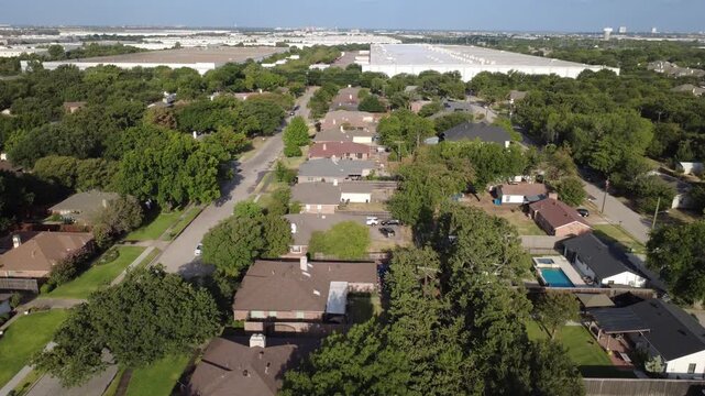 Dense tree canopies border single family neighborhoods near Old Town across DFW area. Massive distribution warehouse rooftops form stark industrial horizon, Coppell, Texas