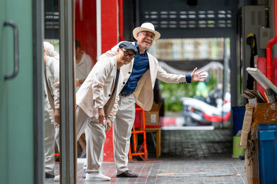 Two senior LGBTQ men in cream suits laugh and lean through doorway, one with straw hat and glasses