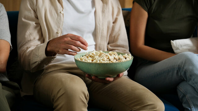 Family members sitting on sofa, sharing a bowl of popcorn and relaxing at home