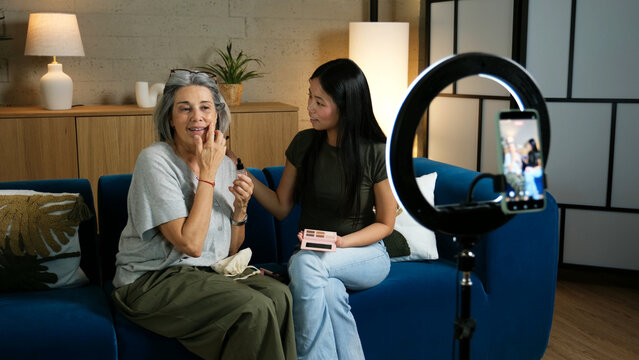 Granddaughter applying makeup on grandmother's face while recording video with smartphone and ring light