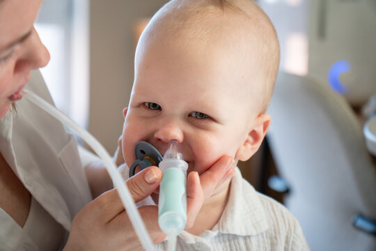 Smiling baby sits with mother as she uses nasal aspirator to clean his nose at home. Infant care, nasal hygiene, parenting, cold relief, caregiving, comfort, health routine, maternal support