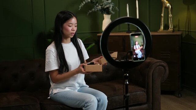 Young asian influencer live streaming and reviewing beauty products using a smartphone and ring light