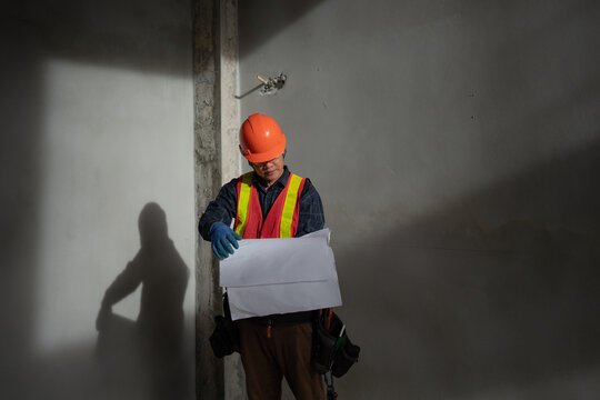 Construction worker wearing a hard hat, reflective safety vest and protective gloves while reviewing architectural plans inside an unfinished building under construction.
