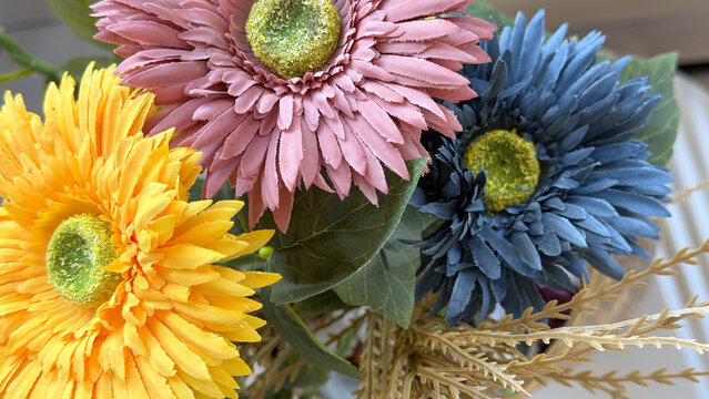 Colorful artificial flower arrangement with yellow, pink and blue blossoms, green leaves and decorative foliage, close-up floral composition for interior decor.