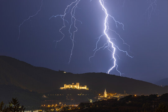 View of the illuminated Rocca Albornoziana fortress atop a hill during a dramatic lightning storm at night in Spoleto, Umbria, Italy.