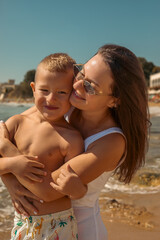 Fototapeta premium Mother embracing her young son on a sunny beach by the sea, enjoying summer vacation, motherhood, Mother's Day, family bonding, relaxation and joyful moments together