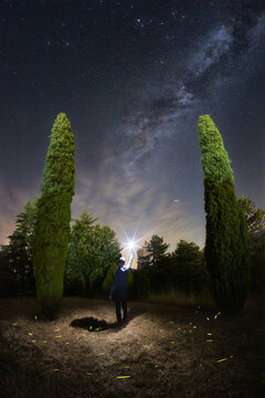 View of a person standing between two tall cypress trees at night, holding a light source towards the Milky Way with fireflies glowing in the field in Fiastra, Marche, Italy.