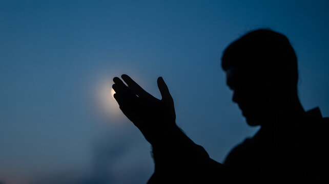 Devout young man praying with his hands held open towards the bright full moon in the dark night sky while reflecting deeply upon his personal faith and spiritual connection during this quiet moment 