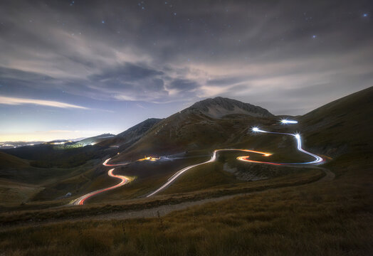 View of the winding mountain road with light trails from vehicles at night beneath a starry sky near Mount Terminillo in Rieti, Lazio, Italy.