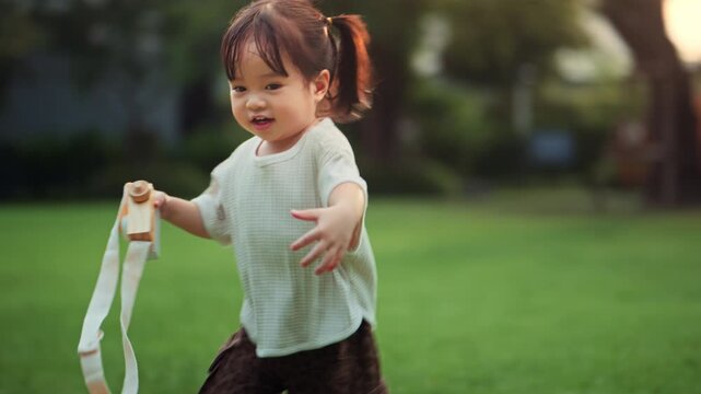 Happy Asian toddler girl running outdoors while holding toy camera, joyful childhood play and active exploration in nature