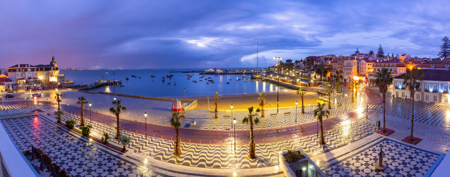 Wide coastal panorama of Cascais with sandy beach, marina, promenade and waterfront buildings at sunrise, Portugal.