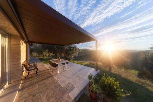View of a patio with outdoor furniture under a pergola overlooking rolling hills and olive trees at sunset in Petriolo, Marche, Italy.