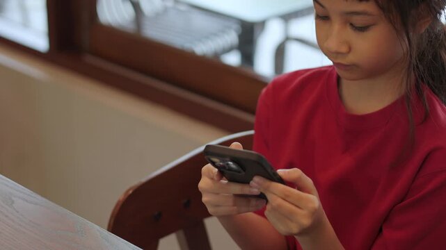 Asian kid using a smartphone at restaurant. Cute Thai girl excitedly watching video or playing games. Shot with selective focus and beautiful copy space for text. Childhood digital lifestyle concept.