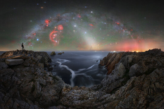 View of a person standing on the rocky cliffs of Pen Hir at night, overlooking the sea under a vibrant Milky Way and colorful nebulae in the night sky, Camaret-sur-Mer, Bretagne, France.