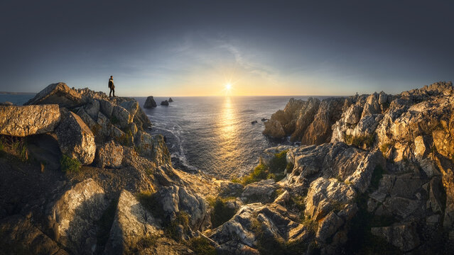 View of a person standing on the rugged cliffs of Pen Hir at sunset, overlooking the Atlantic Ocean with jagged rock formations in Camaret-sur-Mer, Bretagne, France.