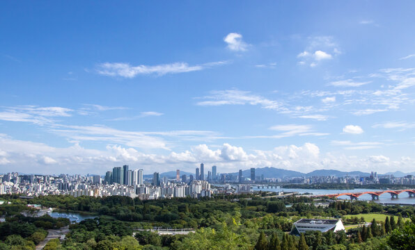 Panoramic aerial view of a large city skyline with skyscrapers, a river bridge, and lush green parks under a vast blue sky
