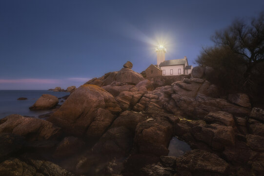 View of Phare De Pontusval lighthouse illuminated at dusk, surrounded by large granite boulders on the rocky coastline in Plouneour-Brignogan-plages, Bretagne, France.