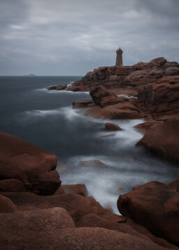 View of Phare de Men Ruz lighthouse on the pink granite coast with crashing waves and cloudy sky in Perros-Guirec, Brittany, France.