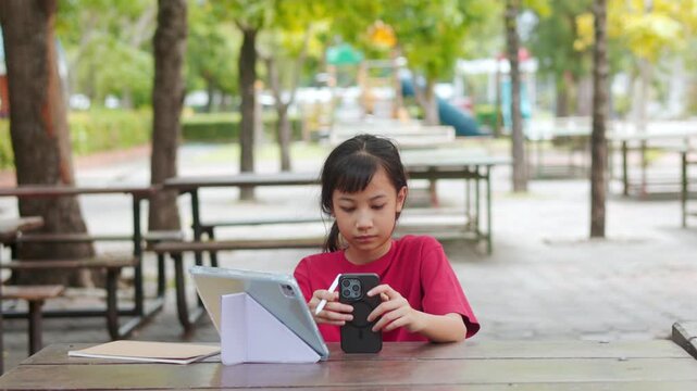 Asian kid doing homework on digital tablet at outdoor campus table. The selective focus shows the student enjoying active e-learning, highlighting modern education technology and tech-savvy childhood.