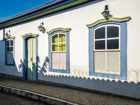 Traditional Brazilian colonial house facade with distinct blue and green wooden windows and a mint-green door. Textured white walls and a cobblestone street under a clear sky. Period wall lamps.