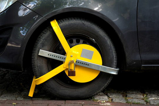 Bright yellow wheel clamp secured around a parked car tire on a paved street, showing vehicle immobilization, parking enforcement, security restriction.