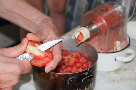 Hands slicing a fresh tomato in a kitchen while preparing homemade tomato sauce, with chopped tomatoes and a manual food grinder in the background.