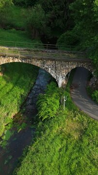 Vertical drone view flying forward towards an ancient stone arch bridge crossing a small river and road in a lush forest