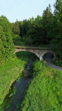 Vertical aerial drone video clip flying forwards over a small river toward an old stone arch bridge in a green forest