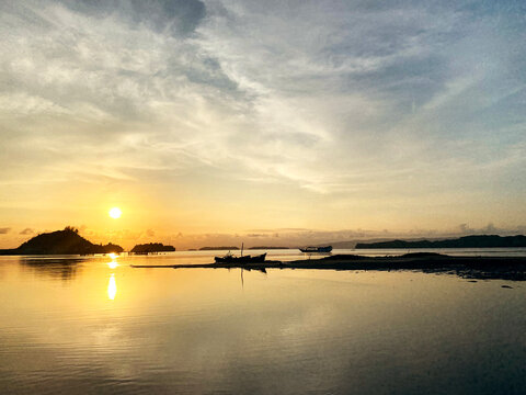 Beautiful tropical sunset over a calm coastal sea, featuring silhouetted islands, traditional boats, distant vessels, and warm golden sunlight reflected across the tranquil water. The peaceful seascap