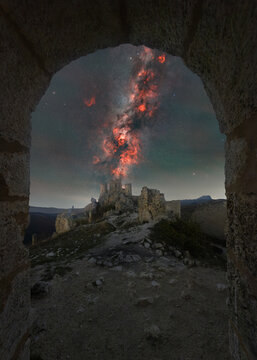 View of the historic Rocca Calascio castle ruins at night, framed by a stone archway under a vibrant, star-filled night sky with a glowing nebula in Calascio, Abruzzo, Italy.
