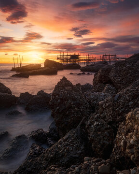 View of the historic Trabocco Spezzacatena fishing structure silhouetted against a vibrant sunset over the rocky coastline in Rocca San Giovanni, Province of Chieti, Italy.