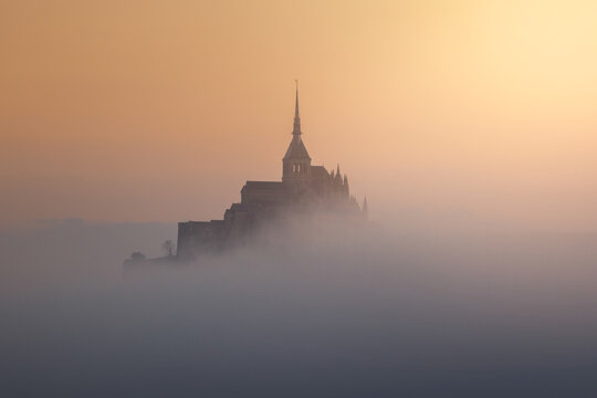 View of the historic Mont Saint-Michel abbey rising above a thick layer of morning fog under a soft golden sunrise light in Mont Saint-Michel, Normandy, France.