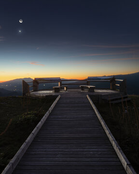 View of the wooden walkway leading to the Rifugio Belvedere viewing platform at twilight with mountains in the background and a crescent moon in the sky in Arquata del Tronto, Marche, Italy.
