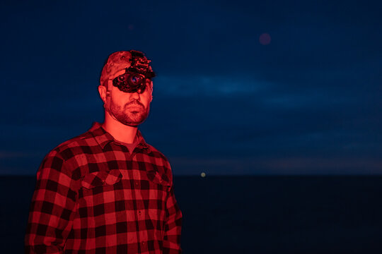 Man in plaid flannel shirt using night vision monocular on skull crusher head rig illuminated by red light at dark night outdoors
