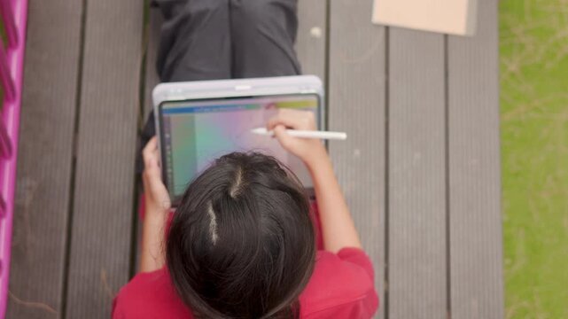 Top view of an Asian kid enjoying a relaxing hobby, drawing digital art on a tablet. Selective focus captures the elementary student spending casual leisure time at an outdoor school.